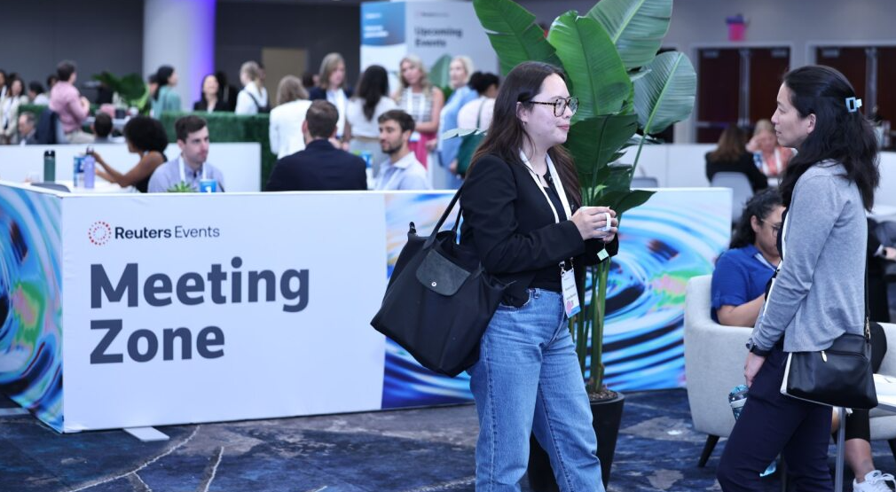 Two women conversing in the "Reuters Events Meeting Zone" at the conference, with other attendees in the background.