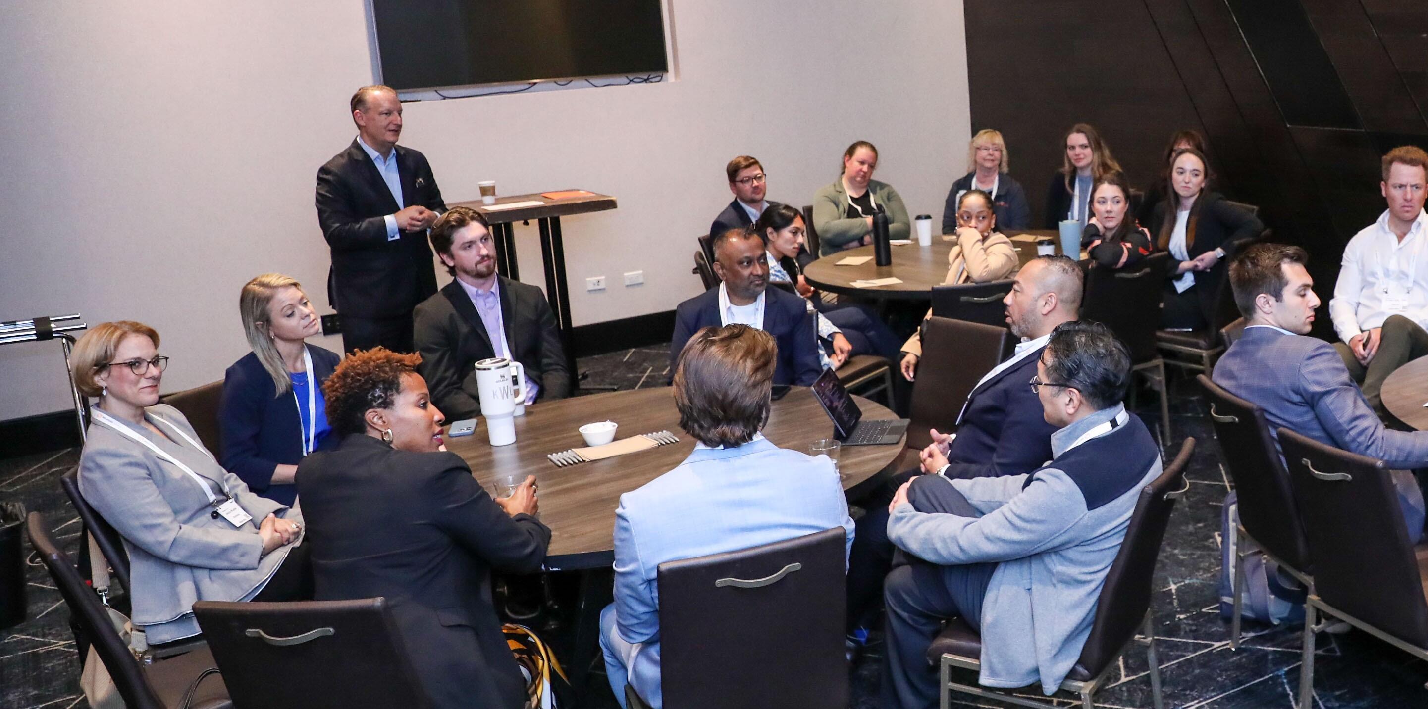 Attendees seated in a semi-circle, listening to a speaker who is standing and gesturing in a meeting room.