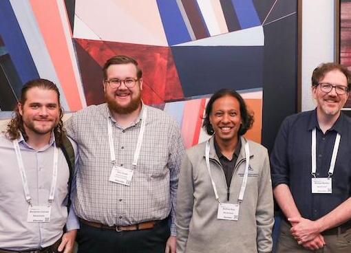 Four men smiling directly at the camera, wearing conference badges, against a colorful backdrop.