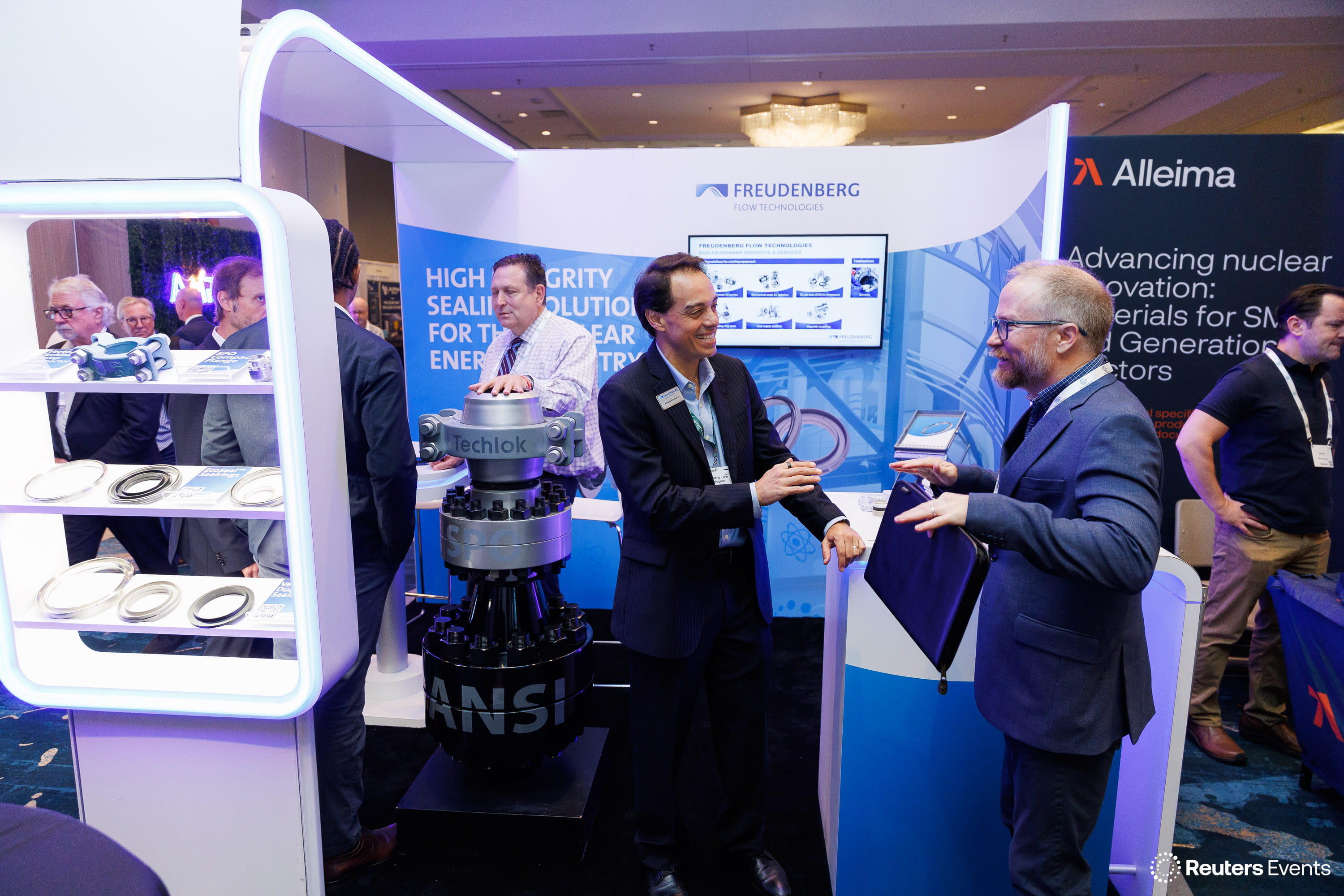 Three men chatting at the Freudenberg Flow Technologies exhibition booth with a Techlok valve display