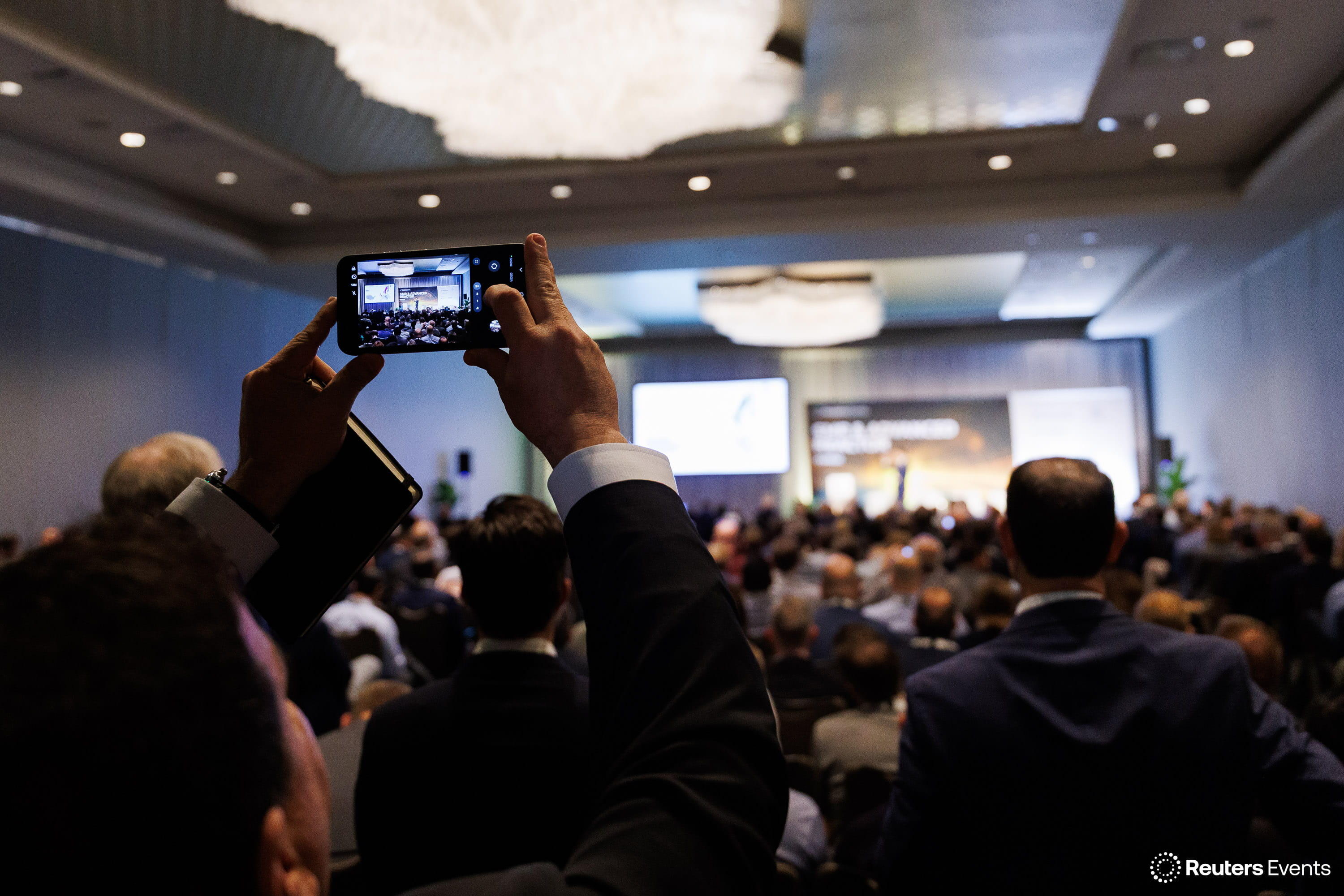 Attendee holds up a smartphone to photograph a keynote in a packed conference ballroom