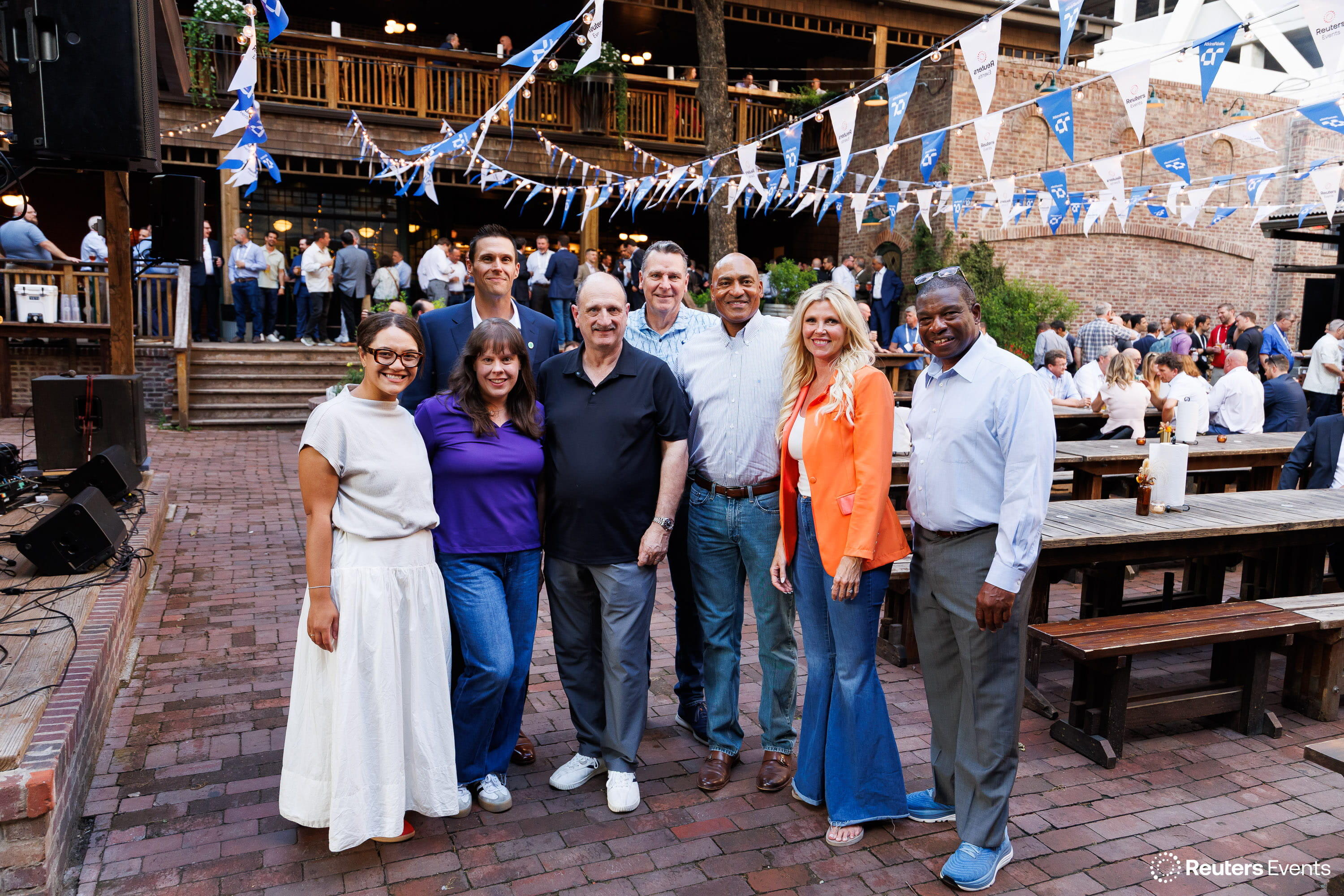 Group of eight attendees posing under Reuters Events bunting at an outdoor evening reception