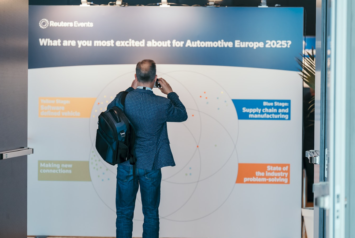 A conference attendee taking a photo of an interactive display asking "What are you most excited about for Automotive Europe 2025?". The display highlights topics like "Software defined vehicle", "Supply chain and manufacturing", "Making new connections", and "State of the industry problem-solving".