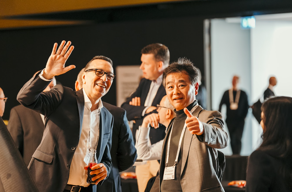 Two men smiling and interacting at the "Automotive Europe" conference. One man waves and holds a drink, the other gives a thumbs up.
