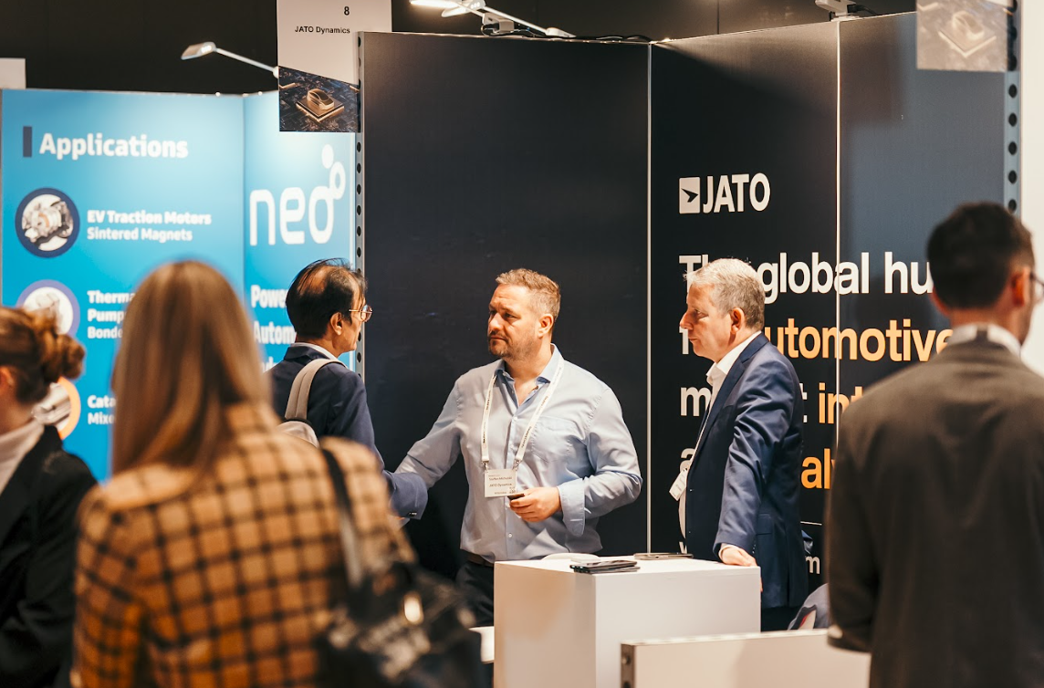Three men converse at a JATO Dynamics booth at the Automotive Europe conference, with "Applications" and "EV Traction Motors" visible on a blue display in the background.