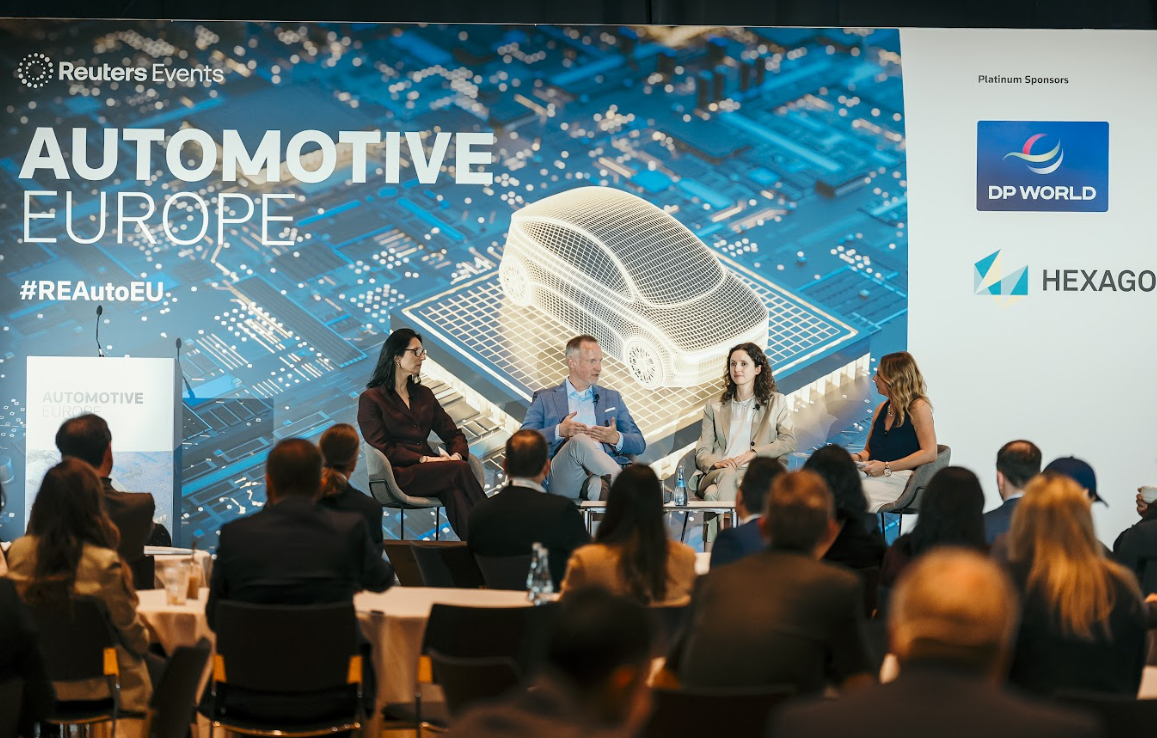 Panel discussion at "Automotive Europe" with four speakers on stage and an audience. The background screen displays "Automotive Europe" and a wireframe car illustration.