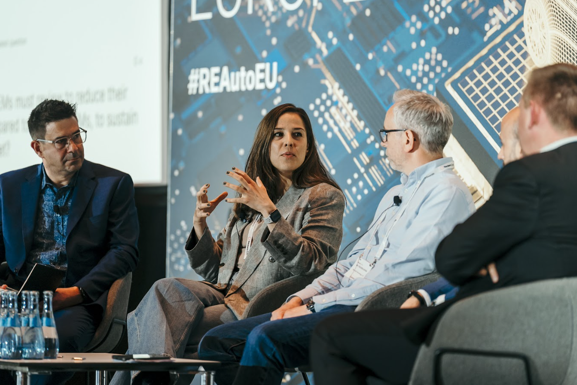 A woman in a grey suit speaks animatedly at the "Automotive Europe" conference, while two men listen. A large screen in the background displays "#REAutoEU".