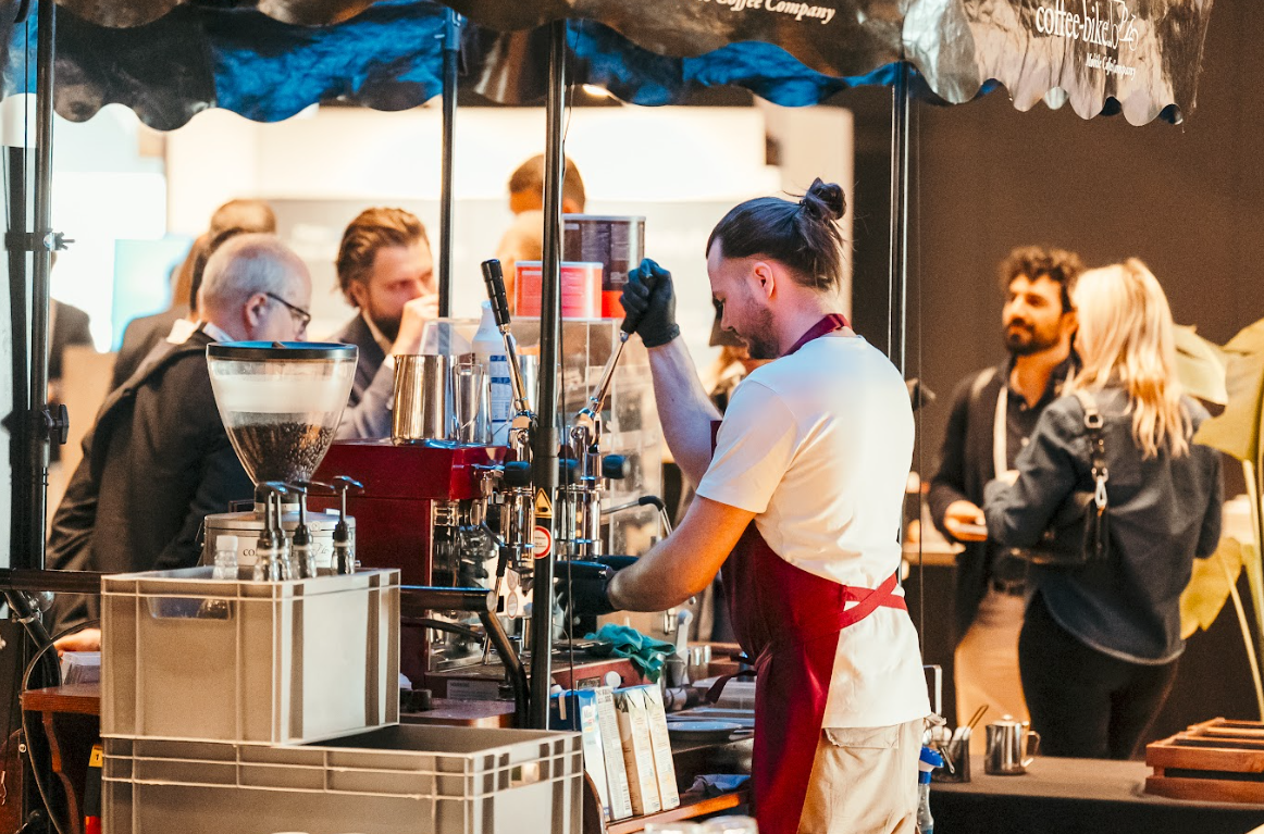 A barista with a man bun and black gloves prepares coffee at a mobile coffee bar, serving attendees at the Automotive Europe conference.
