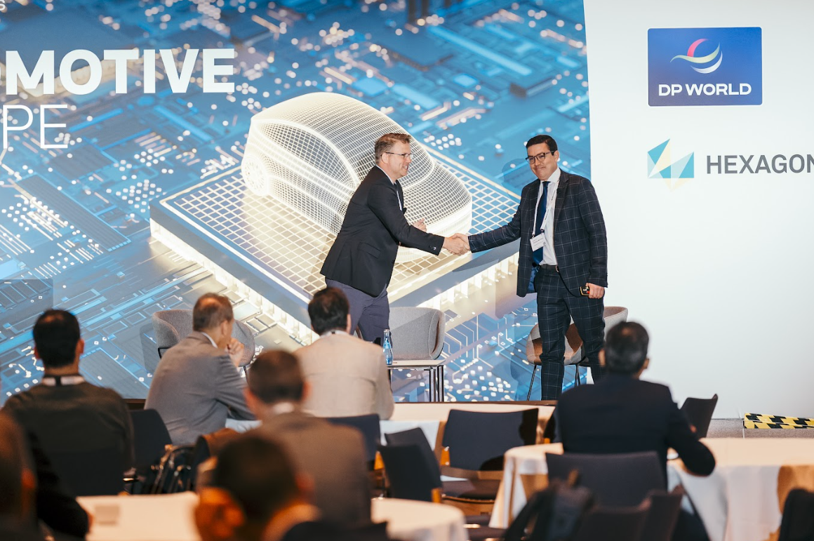 Two men shaking hands on stage at the Automotive Europe conference, with a digital automotive design displayed on a large screen and sponsor logos for DP World and Hexagon.