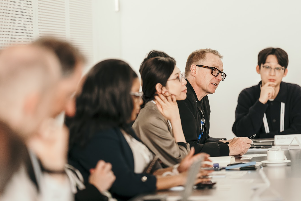 Attendees listening attentively at a conference table, with one man speaking and holding a pen.