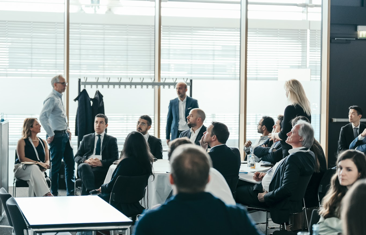 Group of conference attendees, some standing, some seated, engaged in a discussion or presentation in a bright room with large windows.