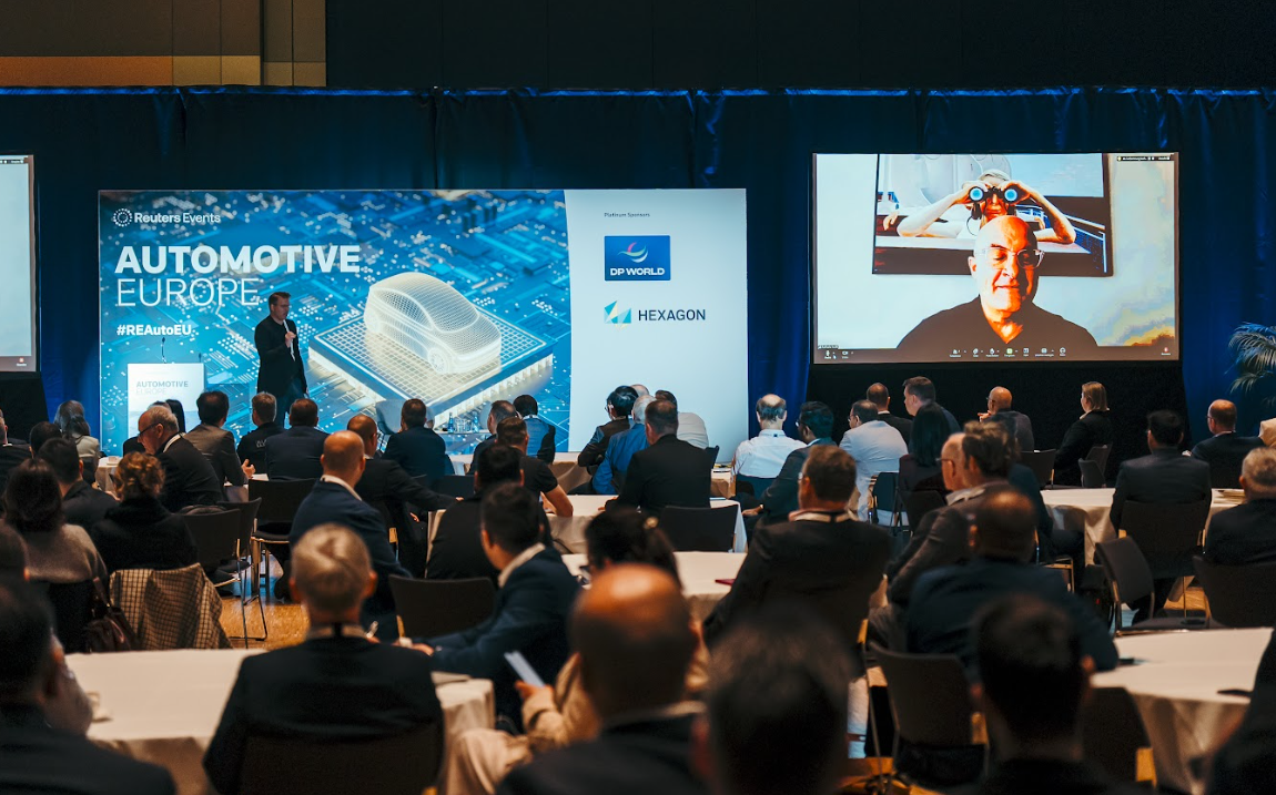 A presenter on stage at "Automotive Europe" conference, addressing an audience, with large screens displaying conference branding, sponsors (DP World, Hexagon), and a remote speaker.