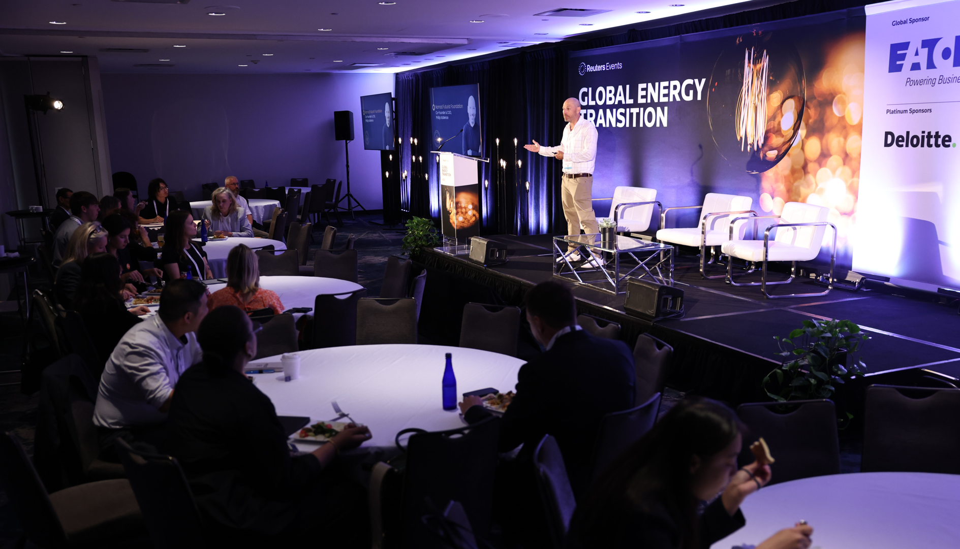 A speaker presents on stage at the "Global Energy Transition" conference by Reuters Events, with attendees seated at tables in the foreground.