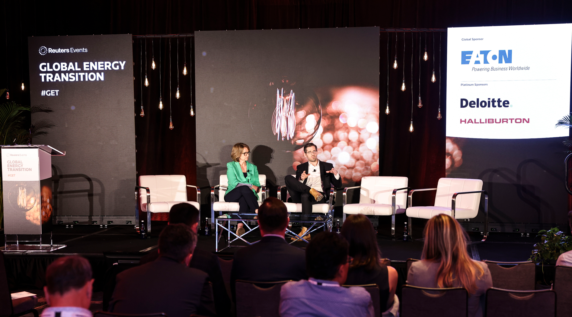Two panelists, a woman and a man, sit on stage discussing "Global Energy Transition" at a Reuters Events conference, with sponsor logos and a large lightbulb graphic displayed on screens behind them.