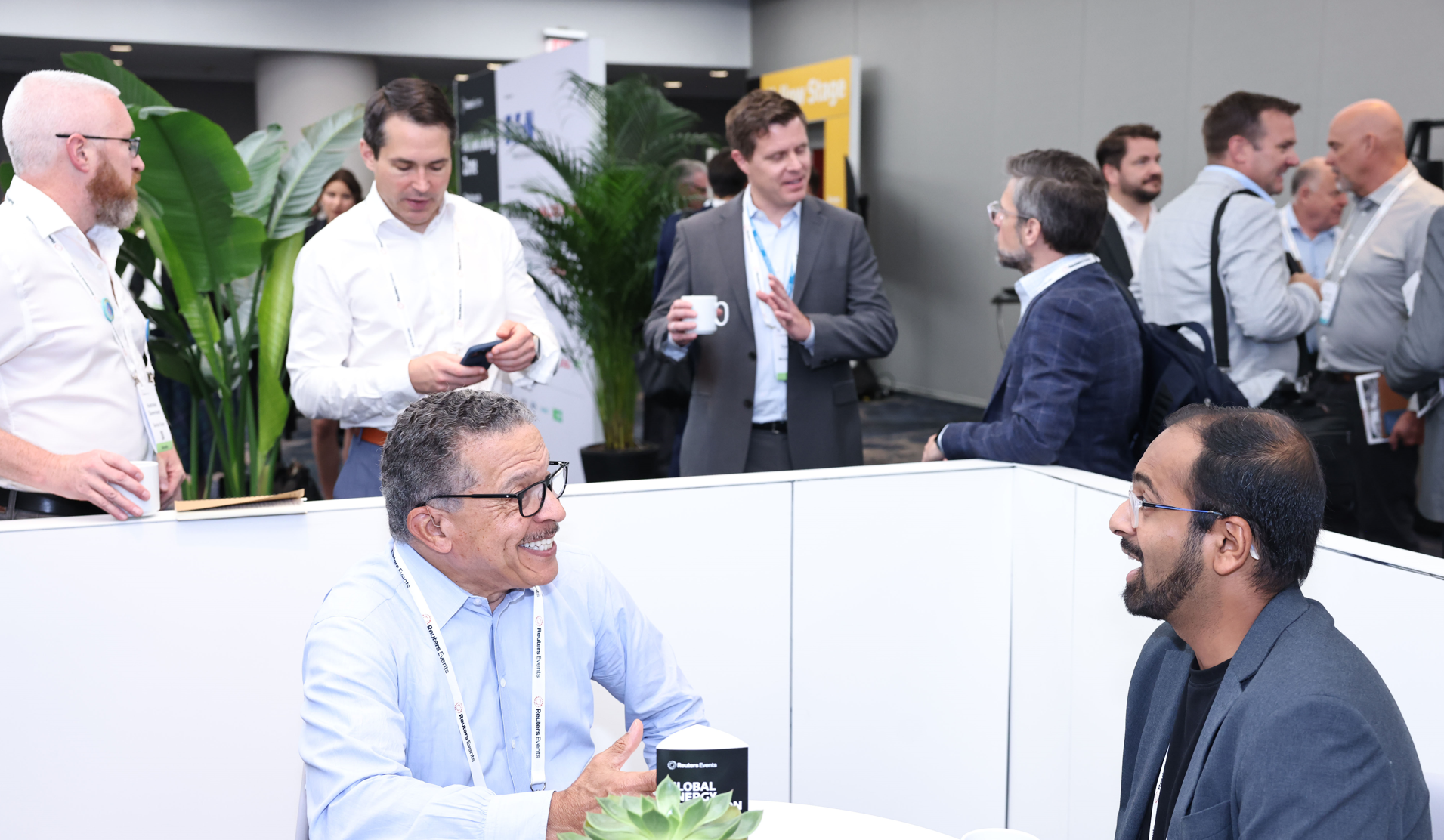 Attendees networking at an event, with two men smiling and conversing in the foreground, and other attendees in the background.