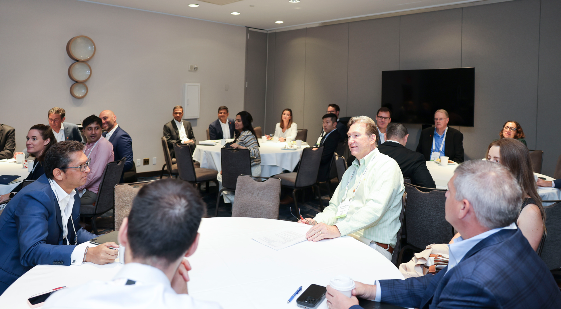Attendees seated at round tables in a conference room, listening to a speaker or presentation.