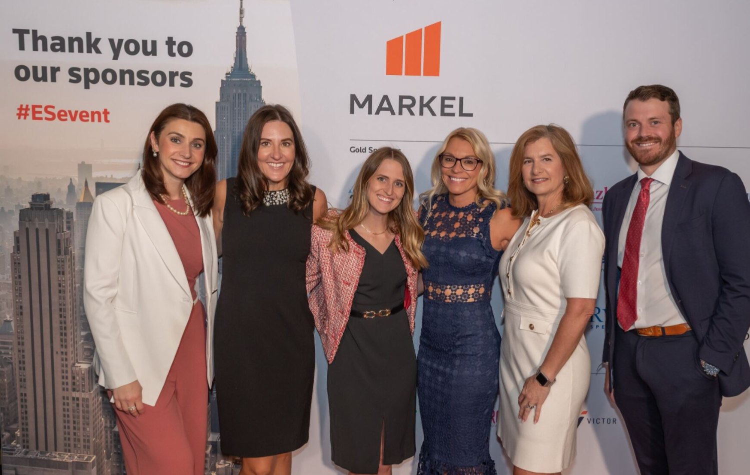 Group of six event attendees posing in front of a sponsor backdrop.