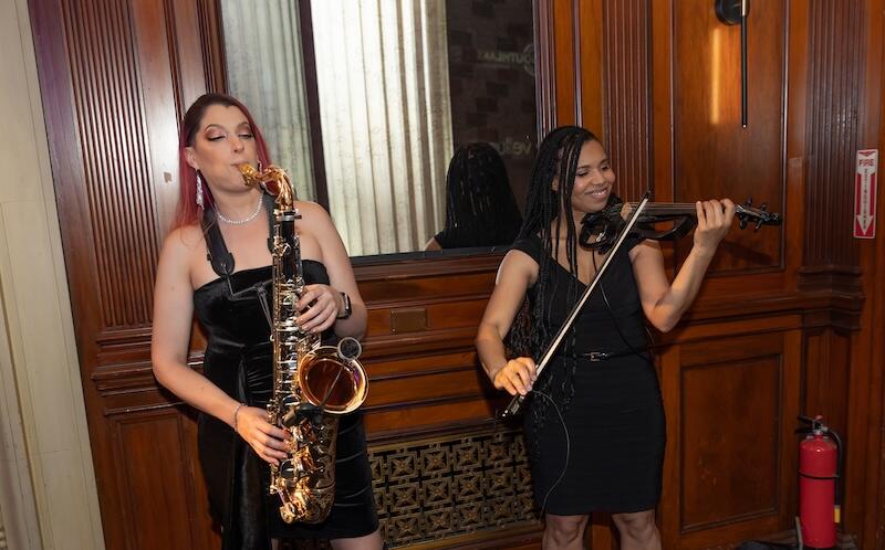 Two female musicians performing indoors, one playing saxophone and the other playing electric violin.