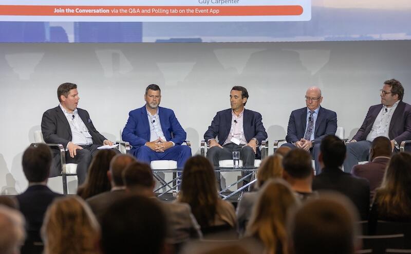 Panel of five speakers seated on stage in front of an audience at a business event.