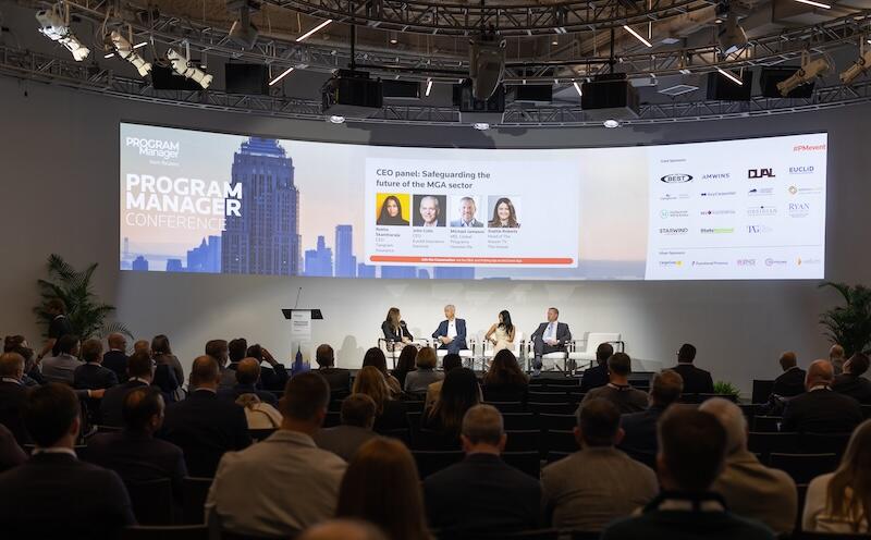 Wide shot of a conference panel on stage with a large "Program Manager Conference" screen in front of a seated audience.