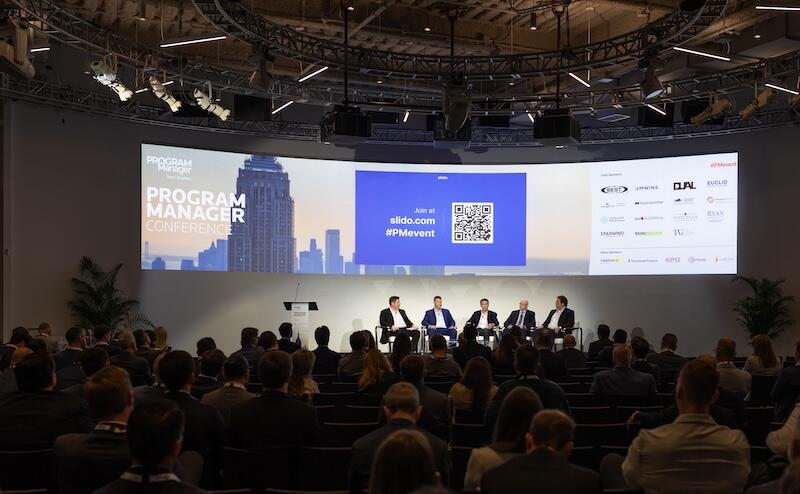 Wide shot of a full conference room watching a panel discussion on stage with a large presentation screen.