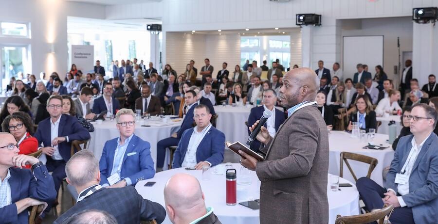 A person standing in a room of people at tables holding a mic  at last years Transform Food & Agriculture USA