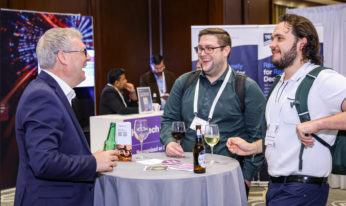 Three men standing around a high-top table with drinks, smiling and engaging in conversation at a conference.