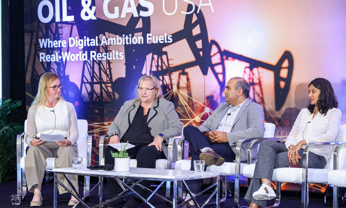 A panel of four speakers, two men and two women, are seated on a stage during a conference. The background features a large screen with text about digital ambition and oil rigs.