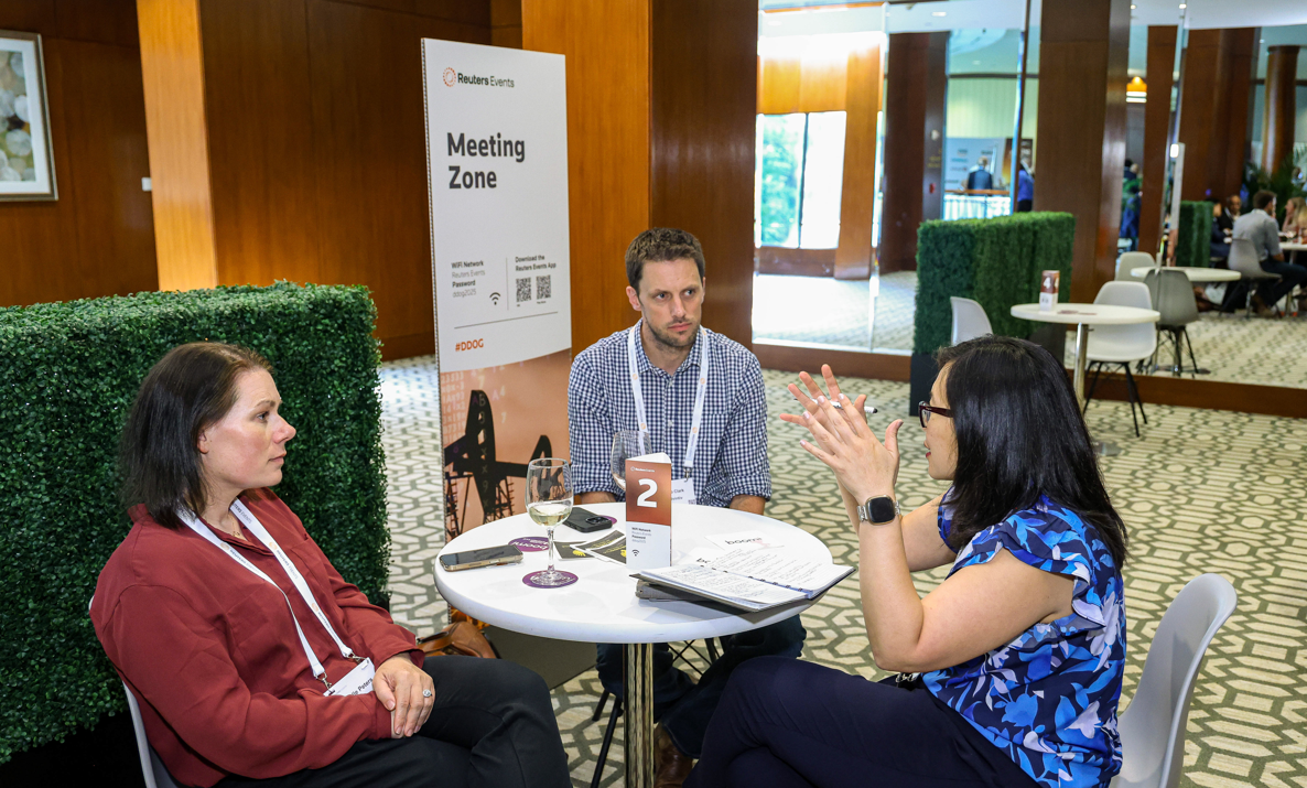Three people are seated around a small, round table at a conference, engaged in conversation. In the background, there's a "Reuters Events Meeting Zone" sign.