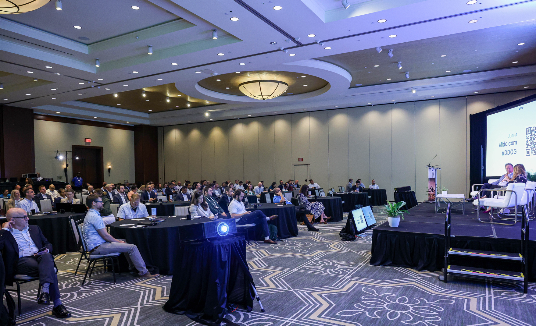 A conference room with an audience seated at tables, watching a presentation on a stage with a large screen displaying "slido.com #DDOG" and a QR code.