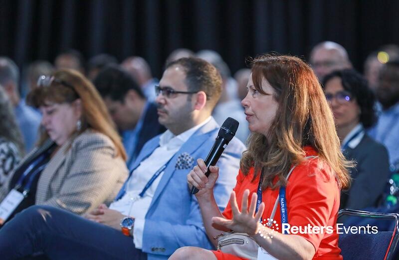 Woman in bright orange top speaking passionately into a handheld microphone during a panel discussion at a Reuters Events conference, with attentive audience members seated around her.
