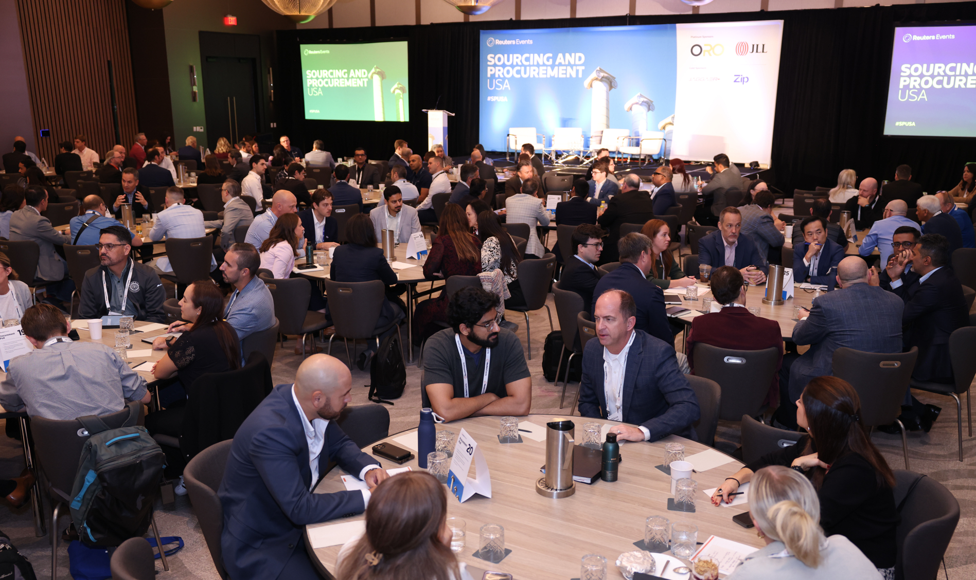 Conference attendees seated at round tables in front of a stage with "Sourcing and Procurement USA" screens.