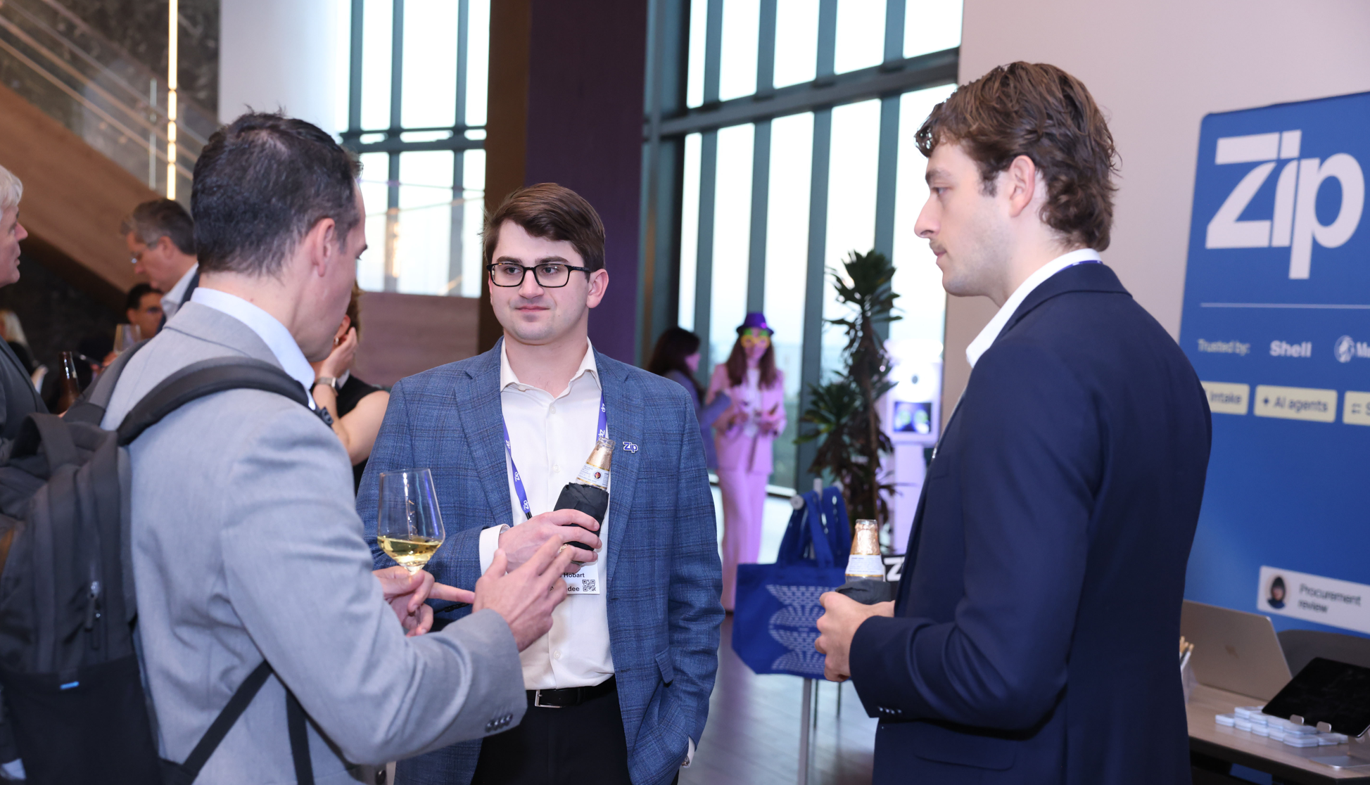 Three conference attendees in suits chat over drinks beside a "Zip" exhibition booth.