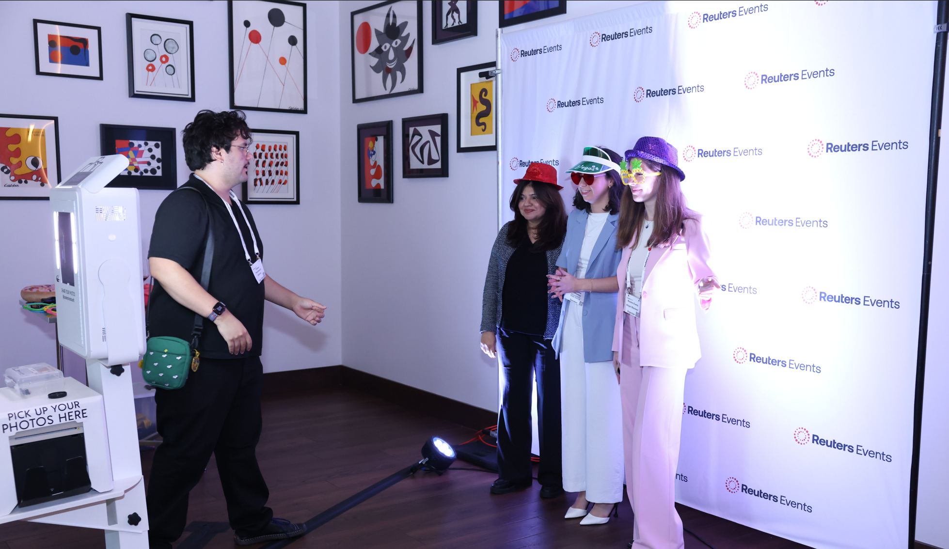Three conference attendees pose in novelty hats and glasses at a Reuters Events photo booth while a staff member adjusts the setup.