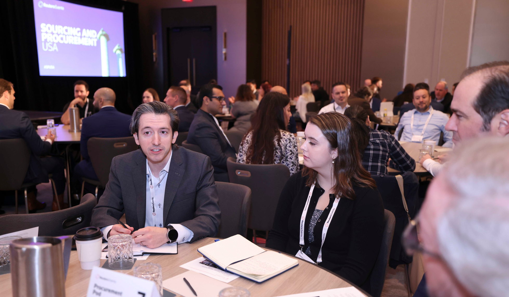 Conference attendees seated at round tables during a networking session, with a "Sourcing and Procurement USA" screen in the background
