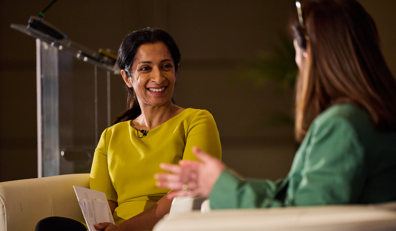 Two speakers seated onstage in conversation, one smiling and holding notes.