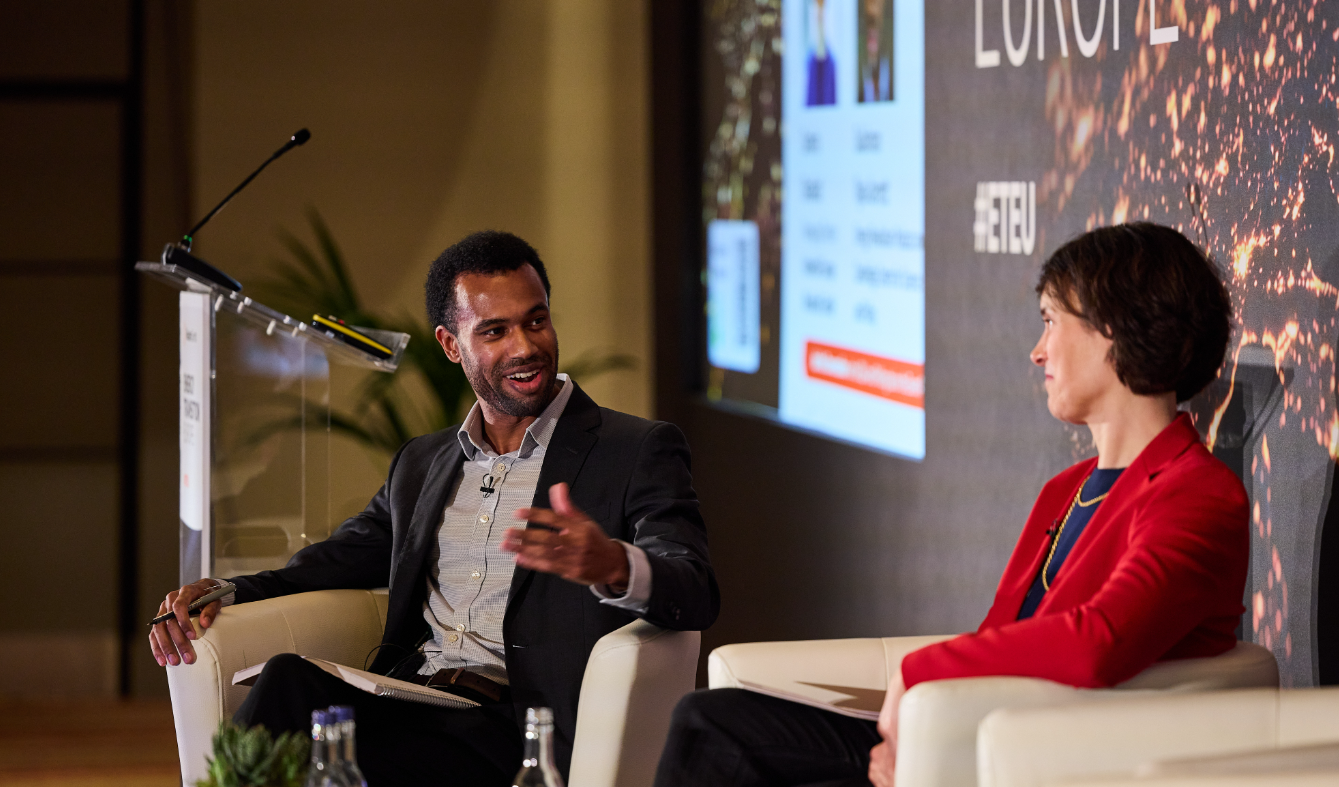Two speakers seated onstage in conversation, with a podium and presentation screen in the background.
