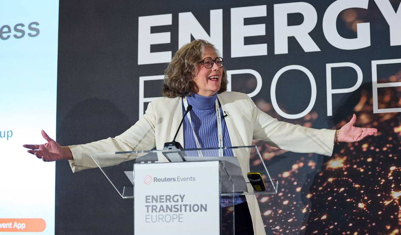 A person is speaking at a clear podium on a conference stage, smiling with arms outstretched as if emphasizing a point. The podium sign reads "Reuters Events" and "Energy Transition Europe," and the backdrop shows large "ENERGY" lettering with a dark, abstract lights pattern. The speaker is wearing glasses, a light-colored blazer over a blue/purple turtleneck, and a lanyard badge, with microphones visible on the lectern.