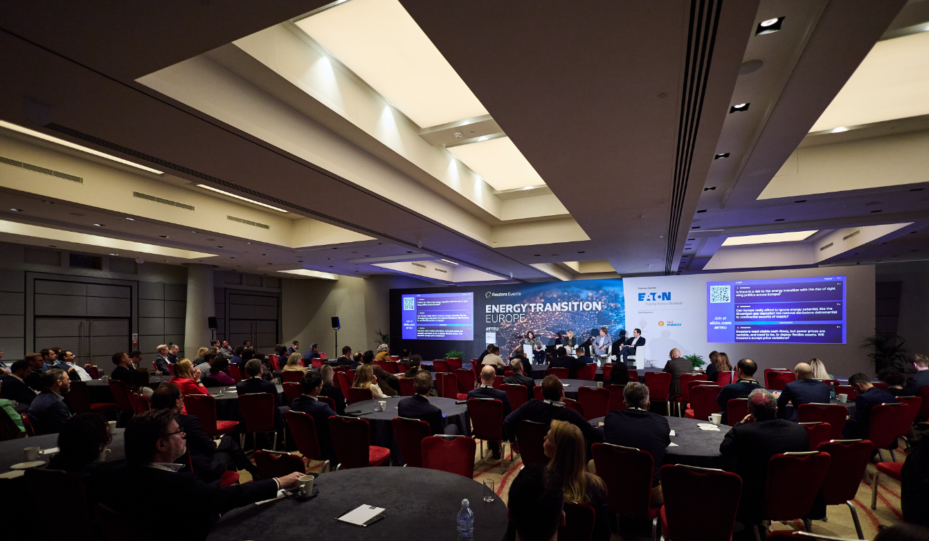 A wide shot of a conference hall during a panel session. Attendees sit at round tables facing a stage with several speakers. Large screens display live questions/QR codes, and the main backdrop reads "Reuters Events – Energy Transition Europe," with sponsor branding (including Eaton).