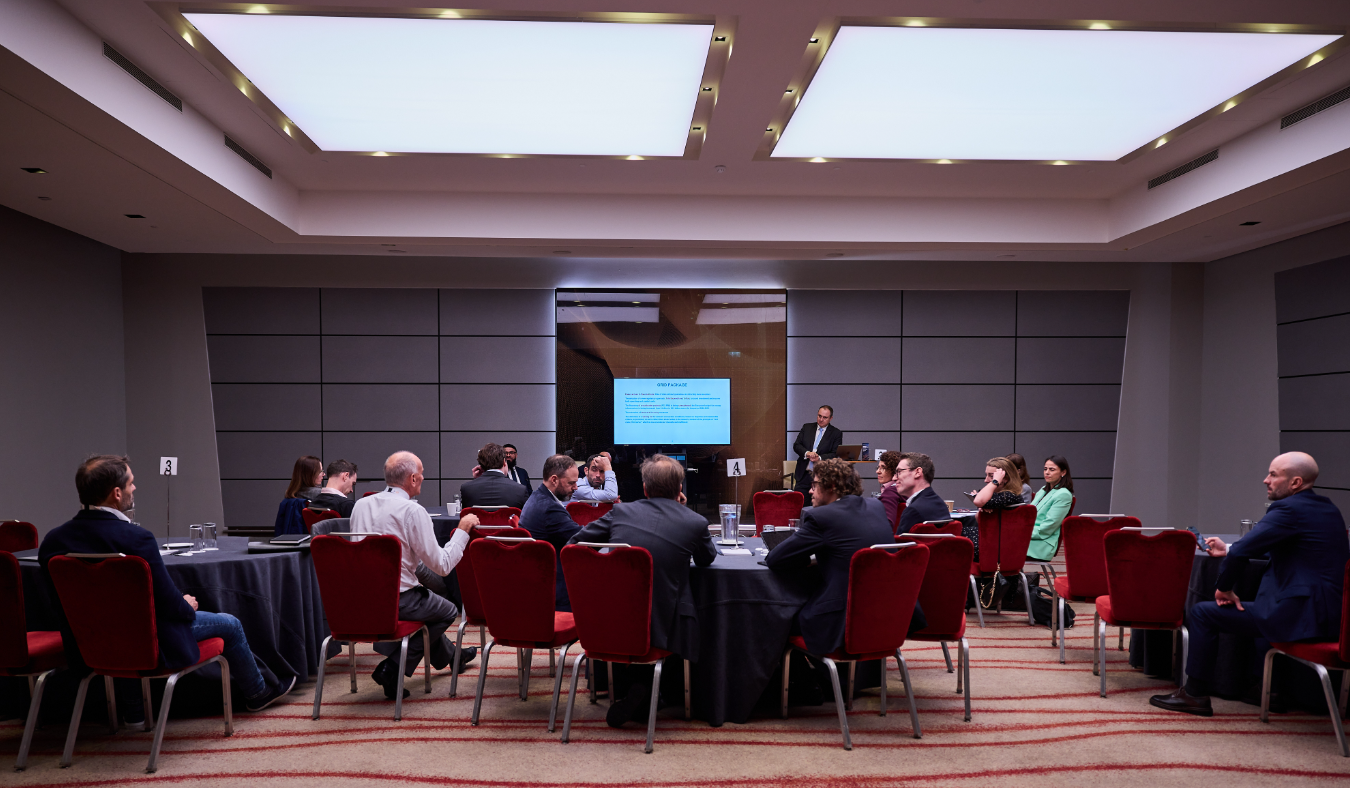 Attendees seated at roundtables during a conference breakout session, with a speaker standing beside a presentation screen.
