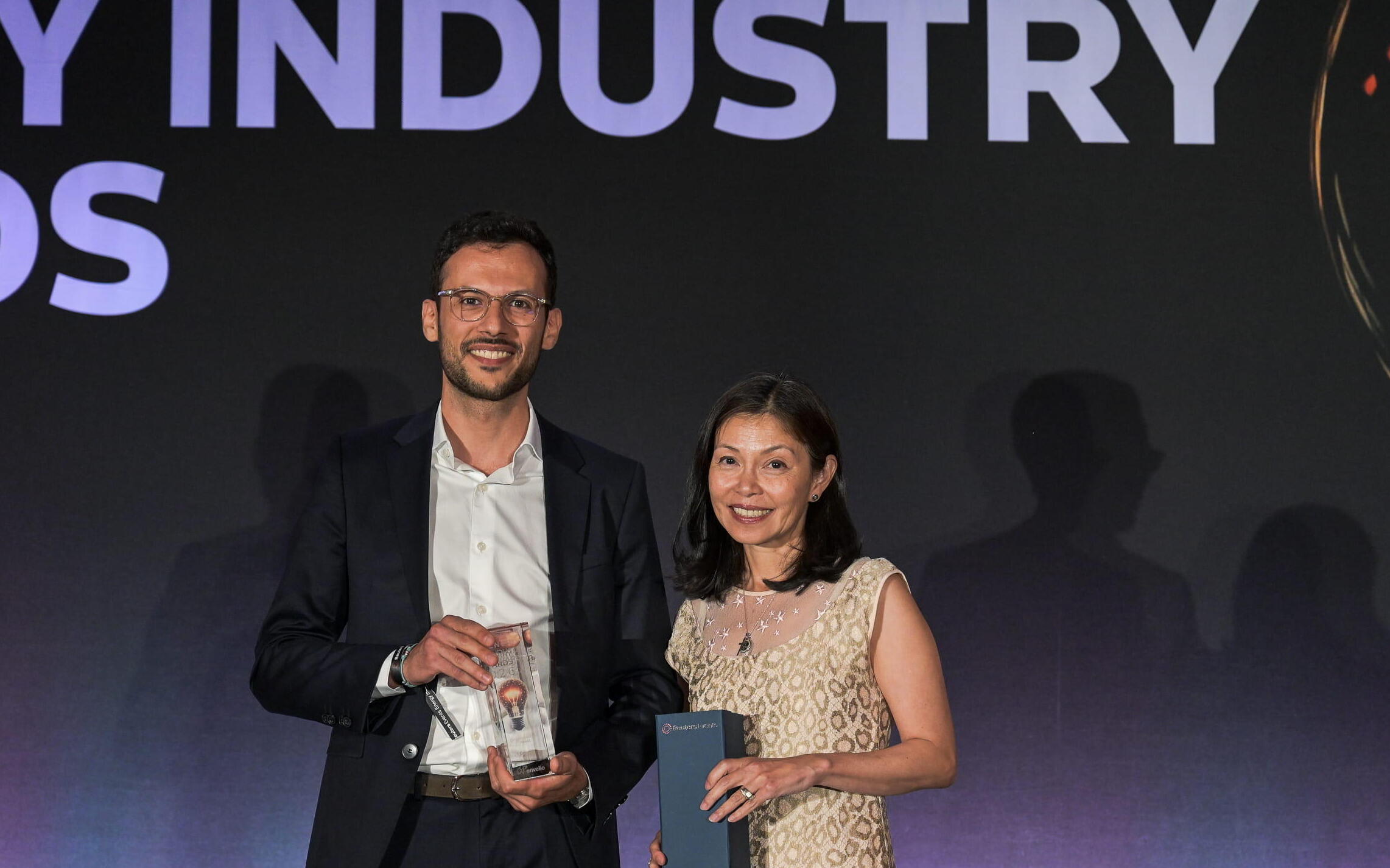 Two award winners smile on stage holding their trophy and gift box at the Energy Industry Awards, with the event name displayed on the backdrop screen