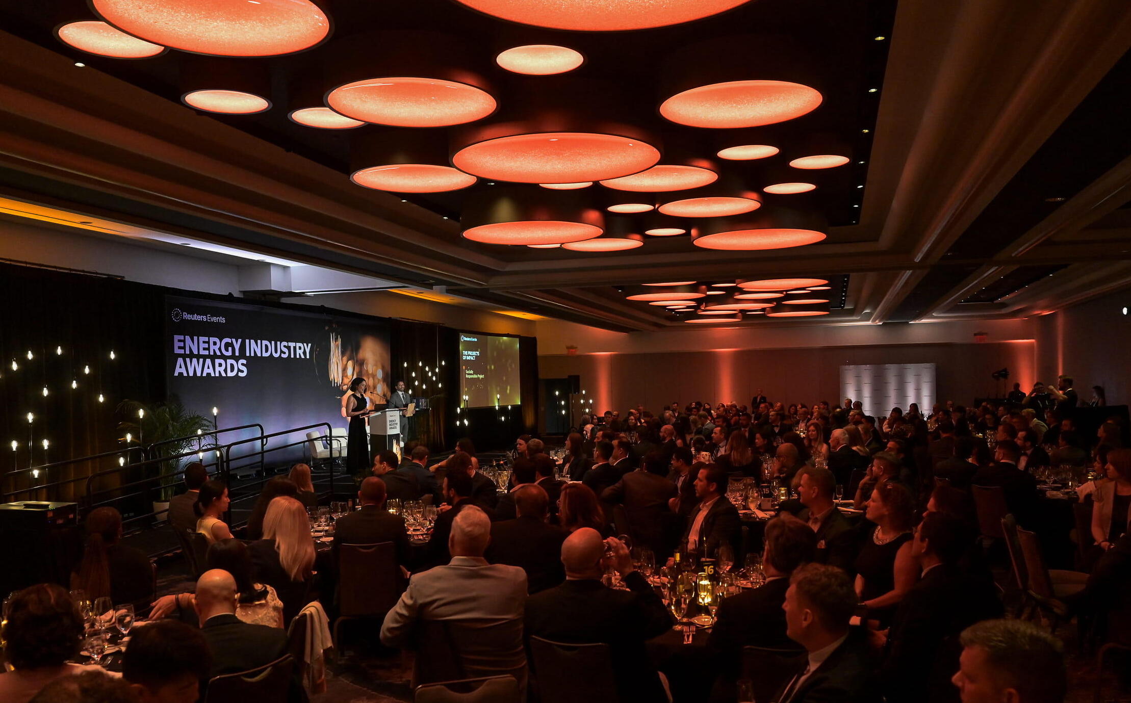 Wide view of the Energy Industry Awards ceremony, showing a seated audience at gala tables in a ballroom with a speaker on stage and 'Energy Industry Awards' displayed on screen