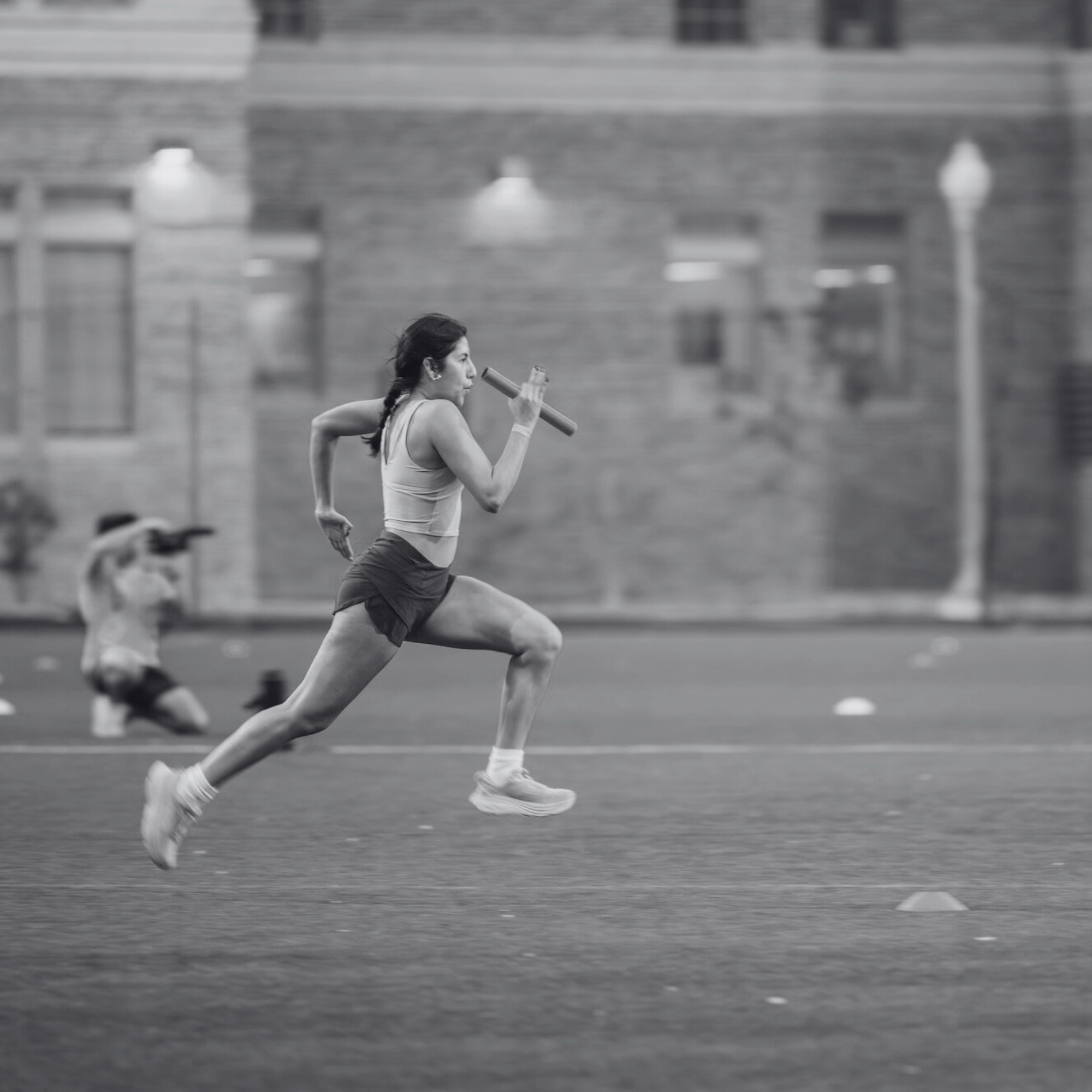A side-profile, black and white action shot of a female runner sprinting across a green field while holding a relay baton. She is captured in mid-stride with intense focus, and a photographer is visible in the blurred background.