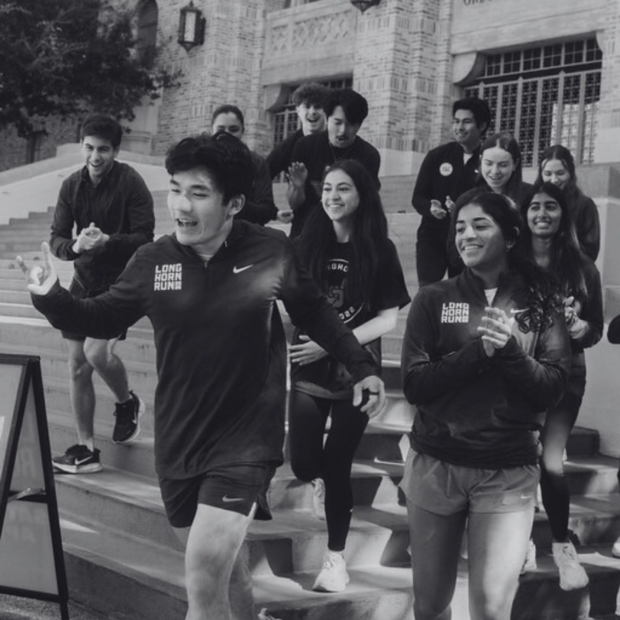 A black and white photo of a large group of runners smiling and cheering as they descend a set of stone stairs outside a university building. Several runners are wearing "Longhorn Run" branded gear.