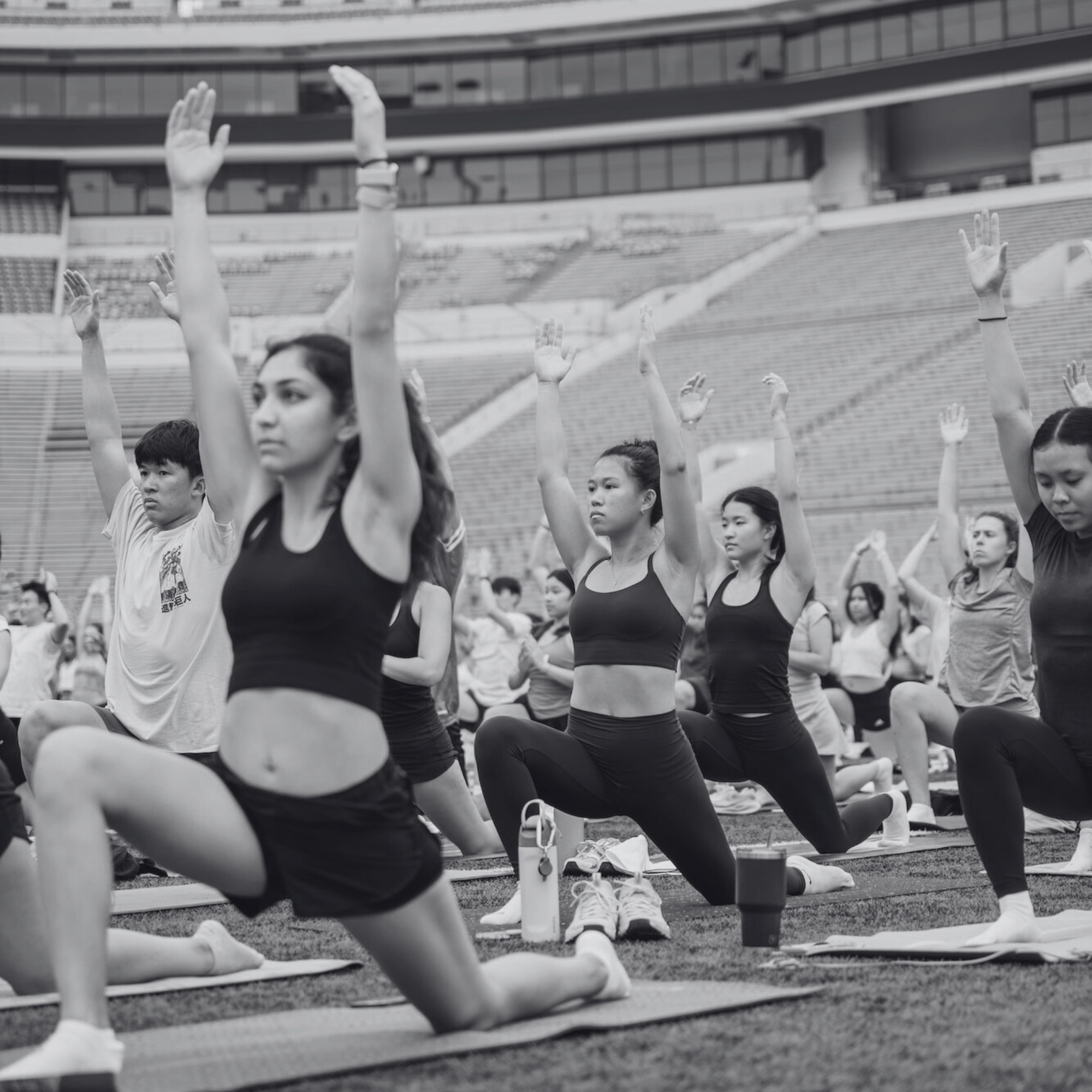 A black and white, low-angle shot of a large group of people practicing a yoga lunge in an outdoor stadium. The participants have their arms raised toward the sky, showing a collective sense of focus and discipline.