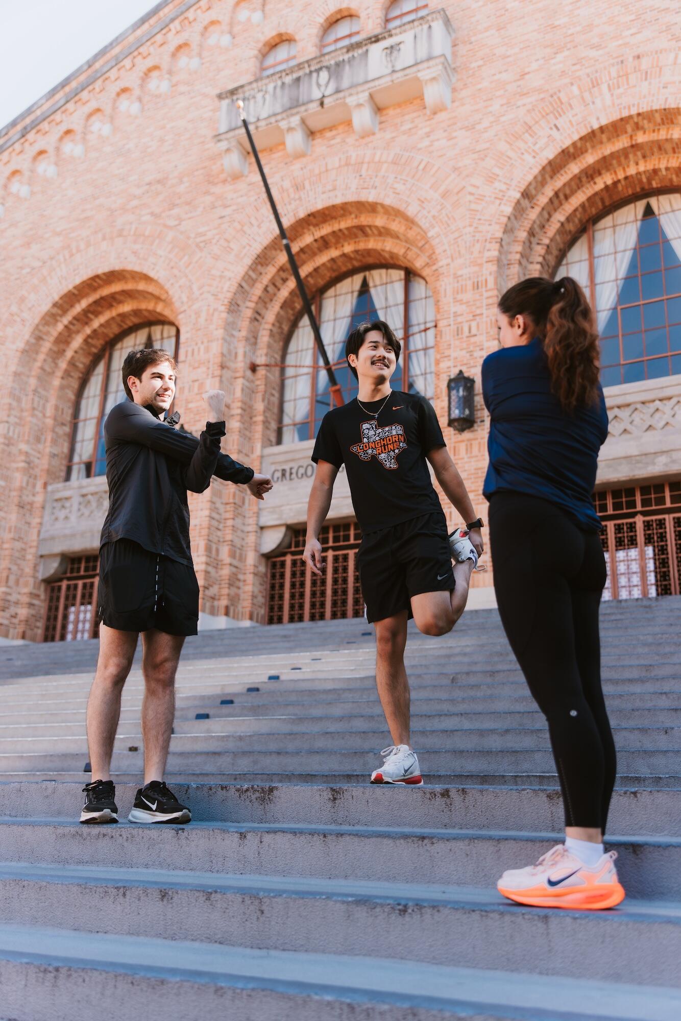 Three runners stretch together on wide outdoor steps in front of a large brick building with arched windows. One person lifts a leg for a quad stretch while the others warm up, wearing athletic clothing and running shoes, suggesting a casual group training or pre-run moment.