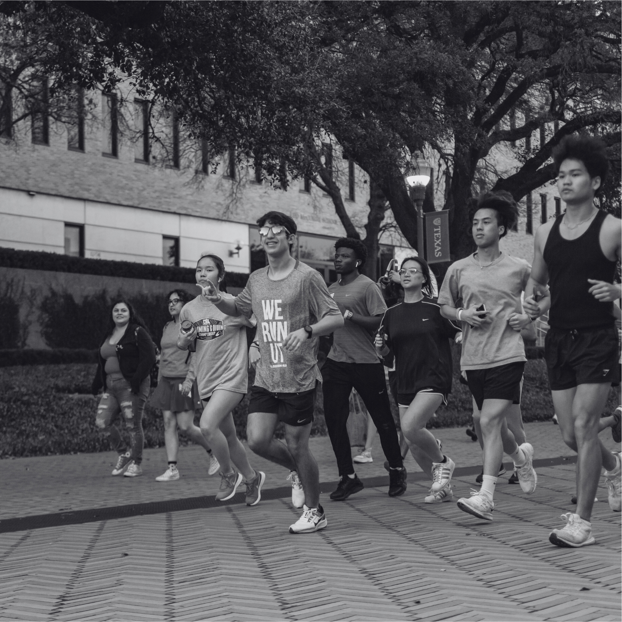 A black and white wide shot of a large group of students jogging together on a brick pathway lined with trees. One runner in the center wears a "We Run UT" t-shirt and reflective sunglasses, smiling at the camera.