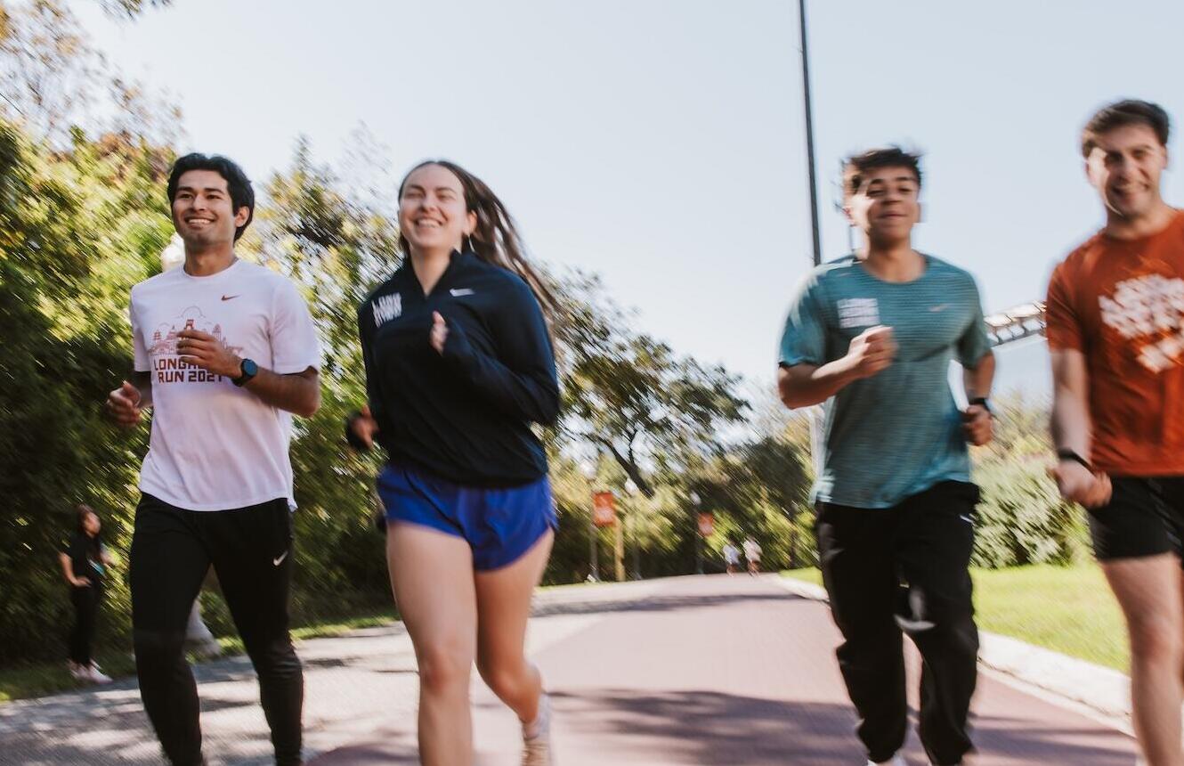 A group of three runners jog together on a paved path outdoors, smiling and mid-stride, surrounded by trees under a clear sky, capturing a lively group run or training session.
