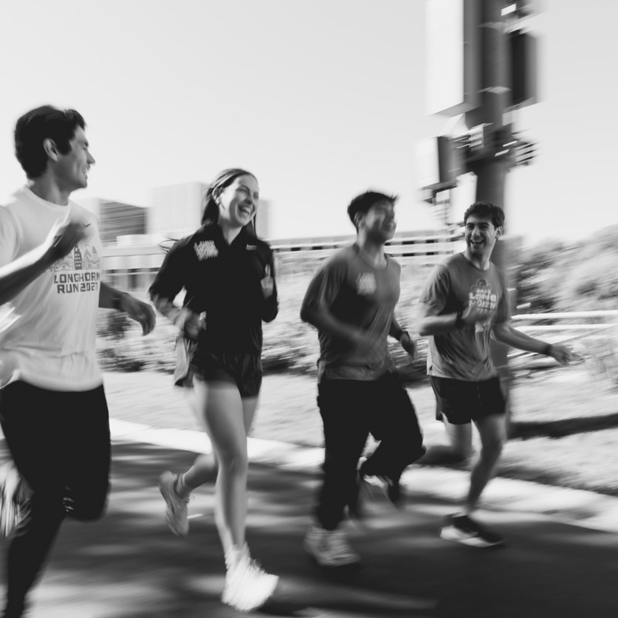 A black and white, motion-blurred photo of four runners laughing and jogging together on an outdoor path. The high-energy, candid shot captures a moment of joy and community during a workout.