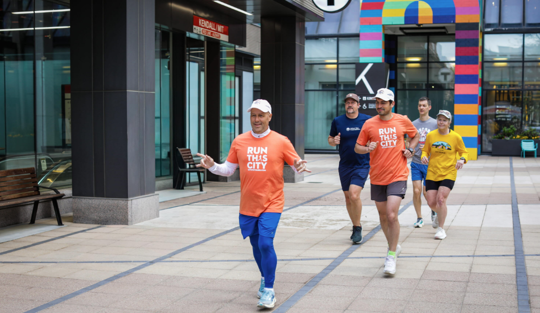 A group of runners in "Run This City" branded t-shirts jog through an urban plaza near Kendall/MIT station at a Reuters Events activity.